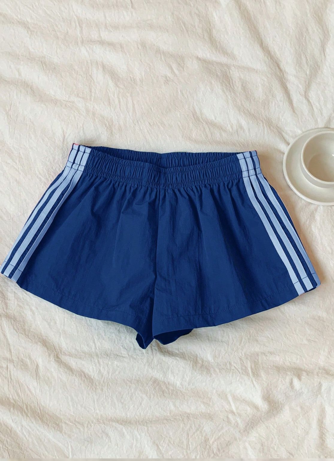 Blue athletic shorts with white three-stripes on the sides, laid flat on a textured off-white fabric background with a small white cup in the top right corner.