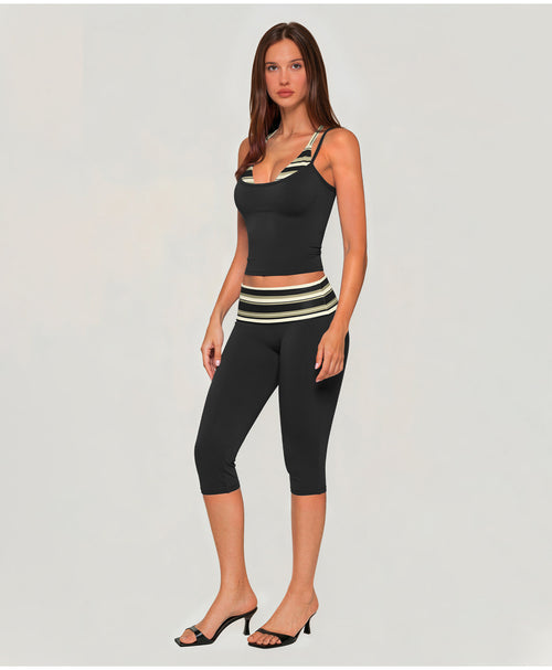 A woman modeling a matching black activewear set consisting of a scoop-neck tank top and high-waisted capri leggings, both featuring a striped waistband and shoulder strap detail in cream and olive green, set against a plain light gray background.