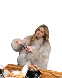 A woman with blonde hair wearing a voluminous light gray fur coat sits at a wooden table holding black sunglasses. In front of her is a cup of latte with chocolate drizzle, french fries, and a knife on a white background.