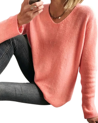 A woman wearing a soft coral pink V-neck knit sweater and dark gray jeans, cropped from the mouth down against a plain white background.