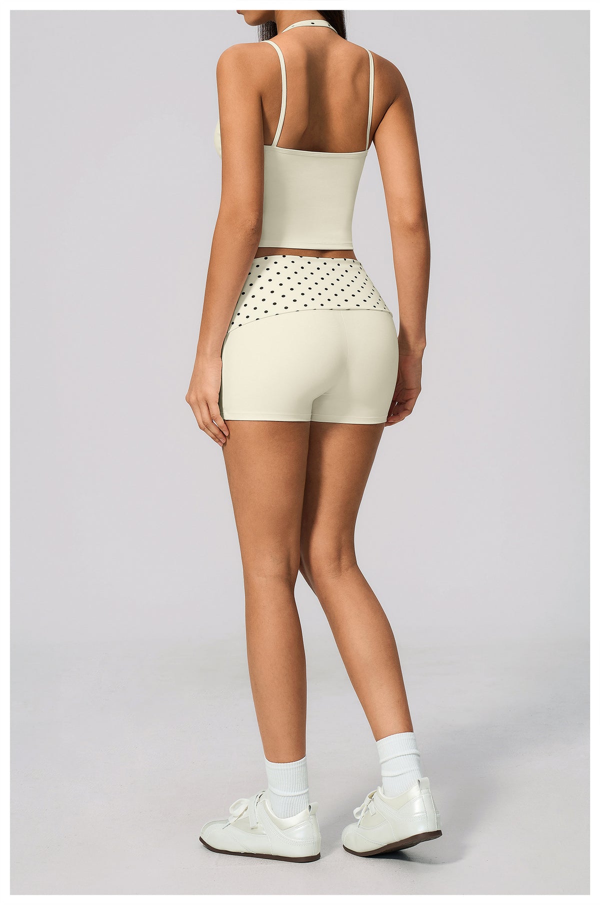 A model shown from the back wearing a matching light beige activewear set consisting of a spaghetti-strap crop top and high-waisted shorts with a dotted pattern on the hip, paired with white socks and sneakers on a light gray background.