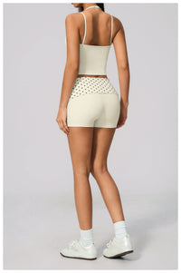 A model shown from the back wearing a matching light beige activewear set consisting of a spaghetti-strap crop top and high-waisted shorts with a dotted pattern on the hip, paired with white socks and sneakers on a light gray background.