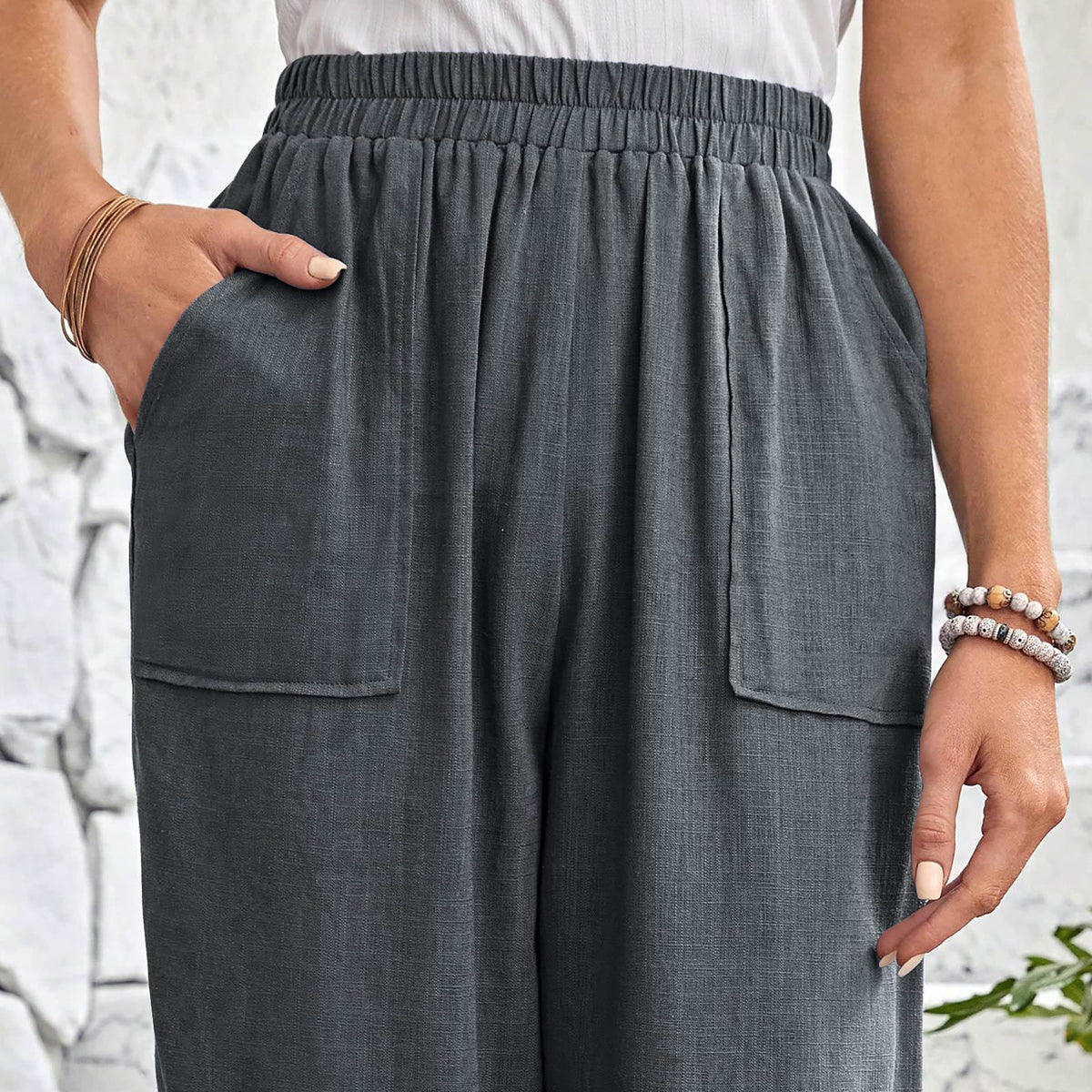 A close-up of a person wearing dark gray wide-leg pants with an elastic waistband, side pockets, and a relaxed drape, shown against a light stone wall background.