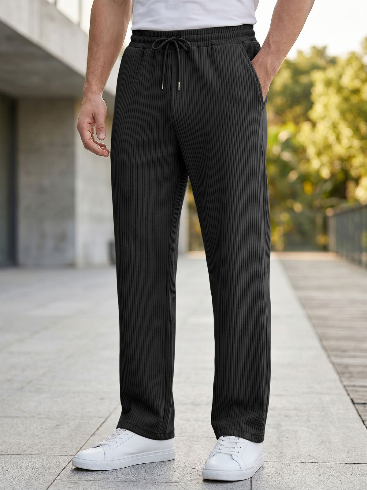 A person wearing dark charcoal gray pinstriped drawstring trousers, styled with a white t-shirt and white sneakers, standing on a paved outdoor walkway.