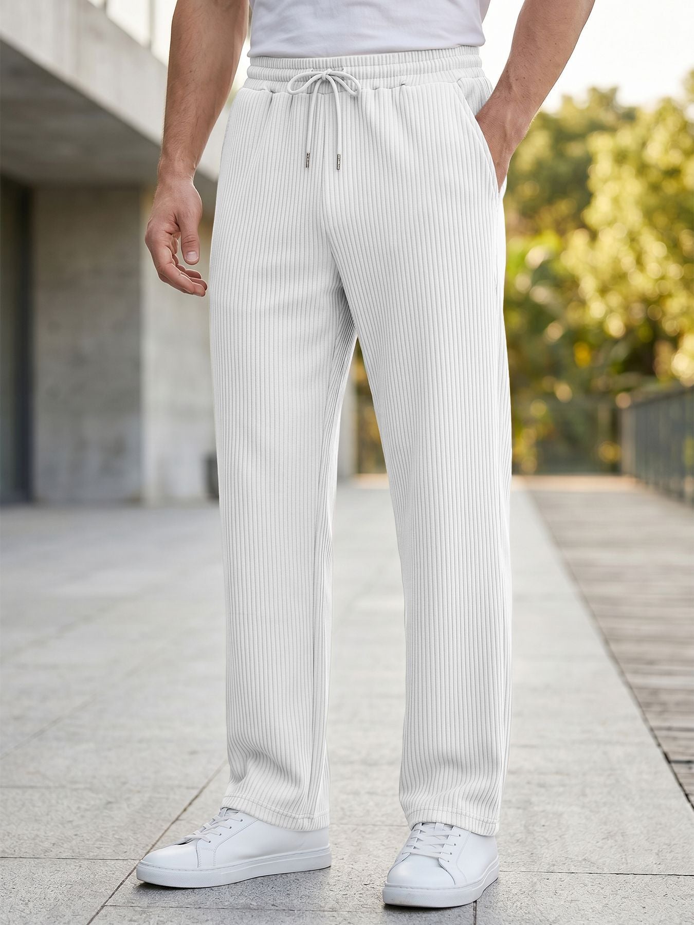 A person wearing white ribbed drawstring trousers and white sneakers, standing outdoors on a paved walkway with blurred greenery and a building in the background.
