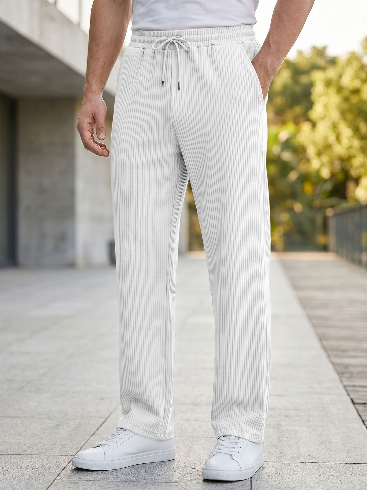 A person wearing white ribbed drawstring trousers and white sneakers, standing outdoors on a paved walkway with blurred greenery and a building in the background.