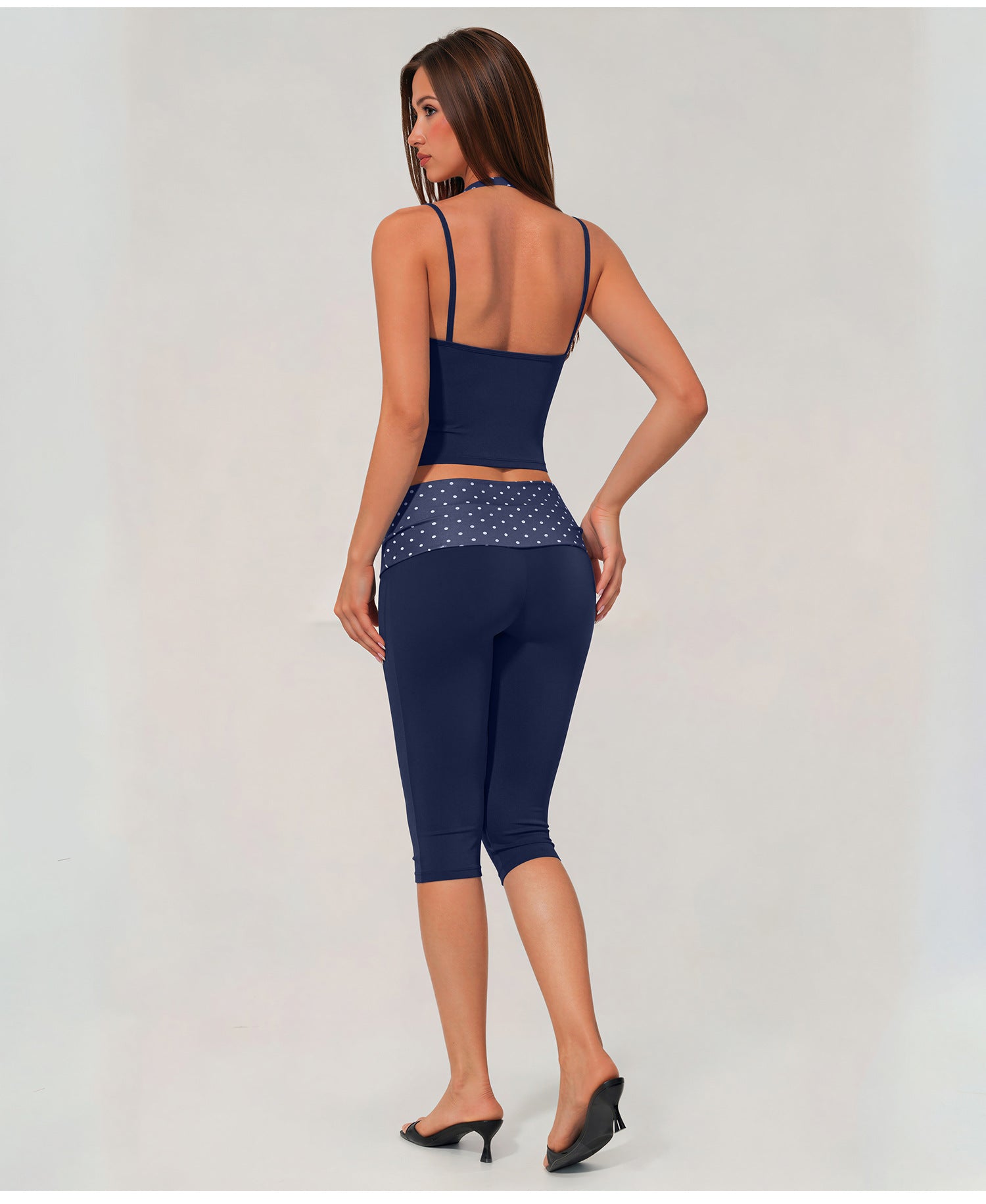 A woman shown from the back wearing a navy blue activewear set consisting of a spaghetti-strap crop top and matching high-waisted capri leggings with a patterned waistband, paired with black heels against a light gray studio background.
