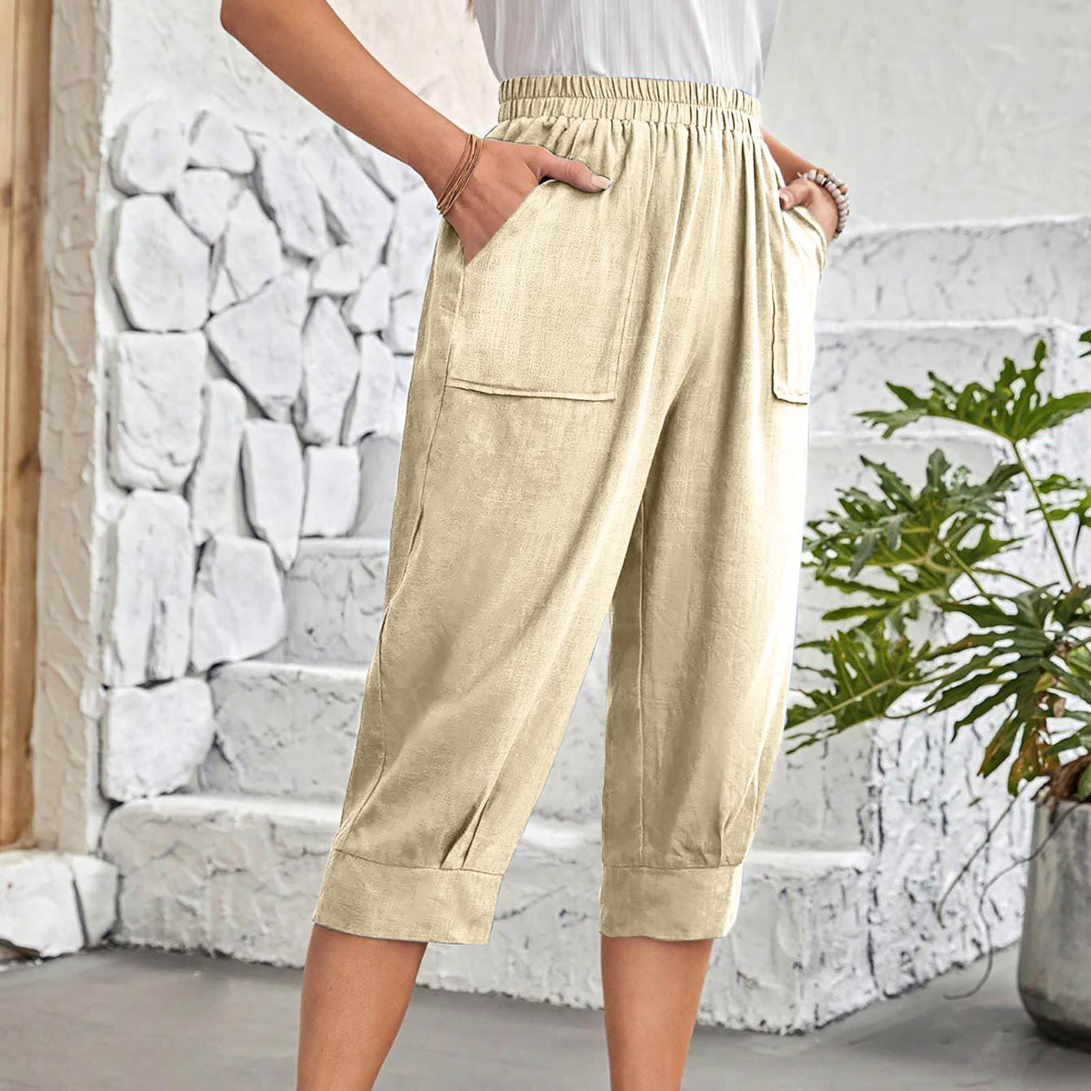 A person wearing light beige cropped pants with an elastic waistband, side pockets, and cuffed hems, standing in front of a white stone wall and potted plant.