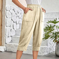 A person wearing light beige cropped pants with an elastic waistband, side pockets, and cuffed hems, standing in front of a white stone wall and potted plant.