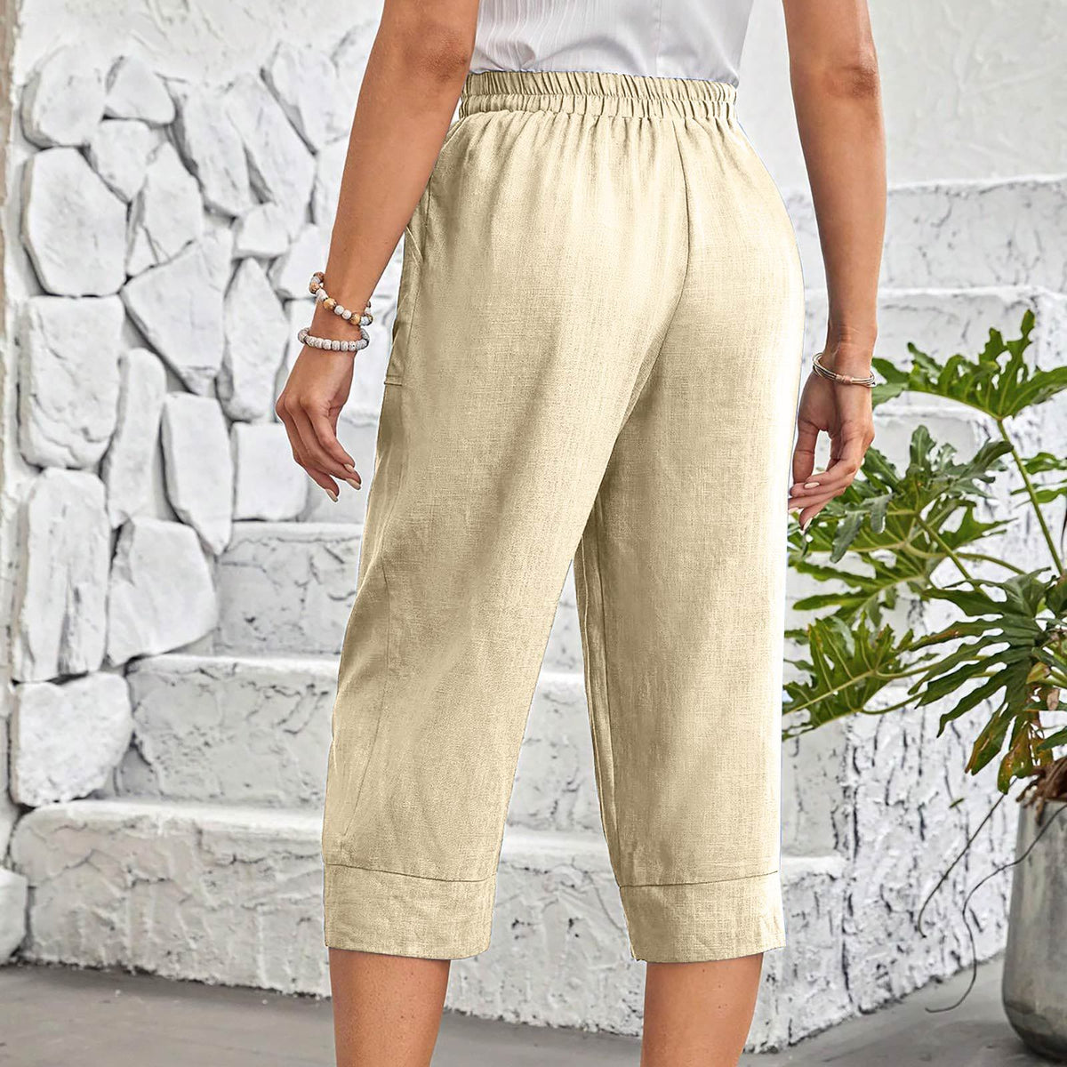 A person wearing light beige cropped pants with an elastic waistband, shown from the back against a white stone wall with a green plant on the right.