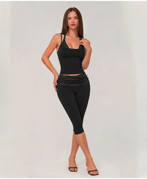 A woman modeling a black two-piece activewear set consisting of a sleeveless camisole top with shoulder straps and matching high-waisted capri leggings with a studded waistband, set against a plain light gray studio background.