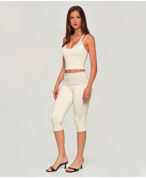 A woman modeling a matching two-piece activewear set consisting of a white scoop-neck crop top with cutout details and white capri leggings with a black polka dot waistband, set against a plain light gray background.