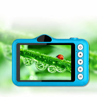 A bright blue children's digital camera with a protective rugged design, featuring a colorful screen showing a ladybug on a dewy green leaf. The camera has four white buttons on the right side and is set against a soft, blurred green background.
