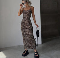 A woman modeling a sleeveless leopard-print maxi dress with thin straps, standing in a minimalist indoor setting with a white wall and concrete floor. She is holding a small black handbag and wearing black platform sandals.
