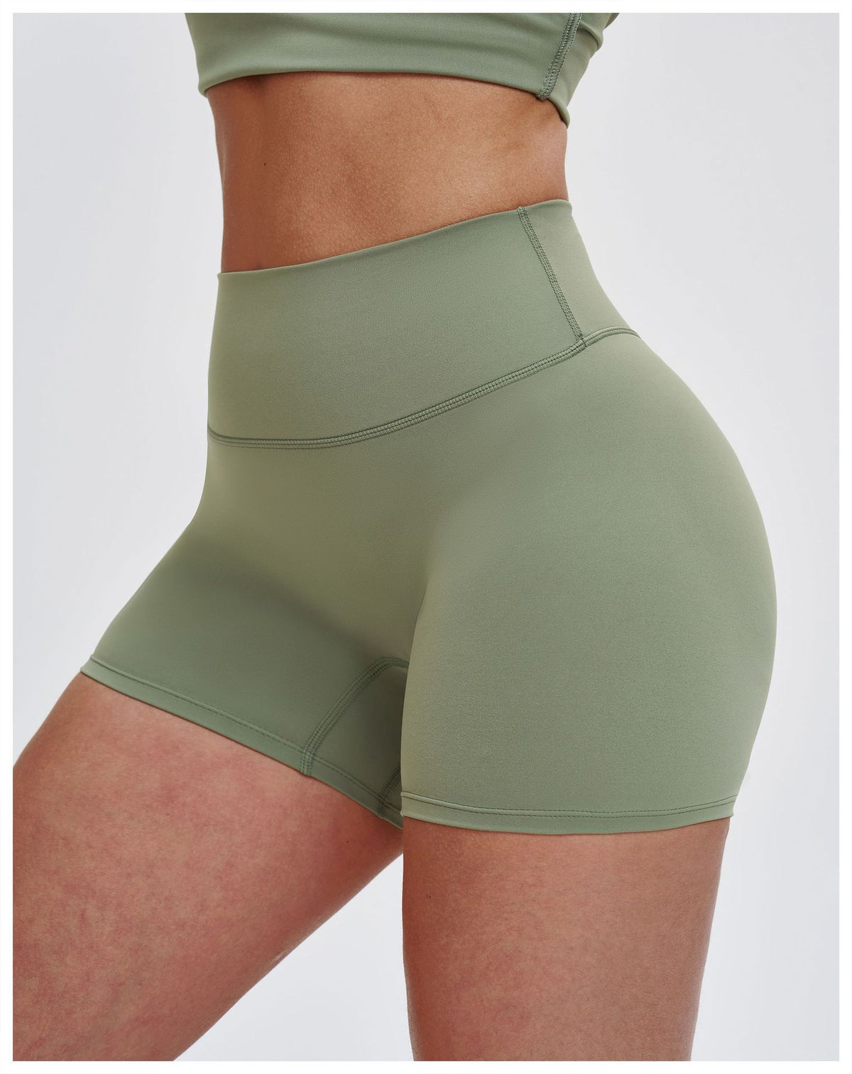 A close-up of a model wearing high-waisted sage green athletic shorts and a matching cropped top against a light gray studio background.