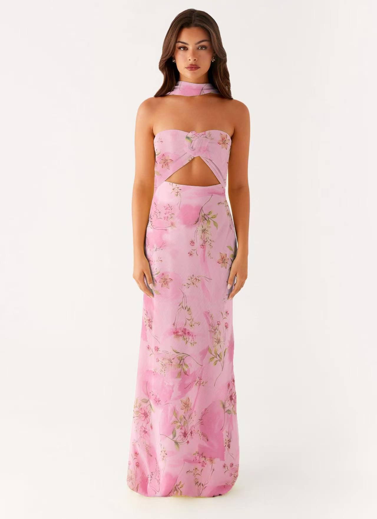 A woman modeling a pink strapless maxi dress with a floral print and a cutout at the waist, set against a plain white studio background.
