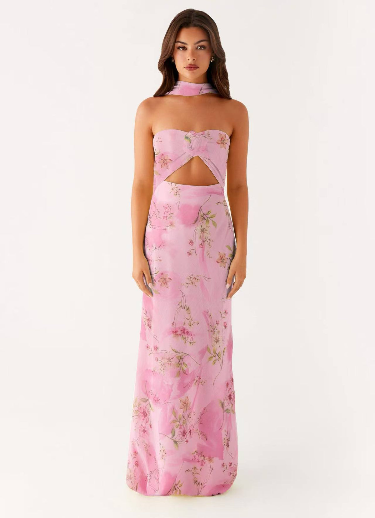 A woman modeling a pink strapless maxi dress with a floral print and a cutout at the waist, set against a plain white studio background.