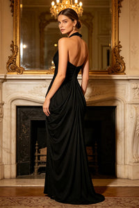 A woman wearing a floor-length black evening gown with a draped back and halter neckline, standing in front of an ornate fireplace and mirror in a luxurious interior.