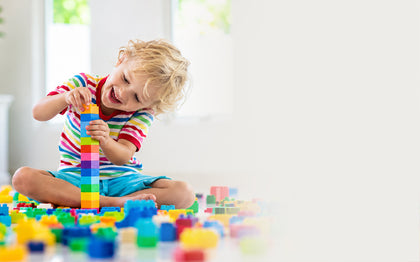 A young child with blonde hair sits on the floor playing with colorful building blocks, holding a tall striped tower. The child wears a rainbow-striped shirt and blue shorts in a bright indoor room.