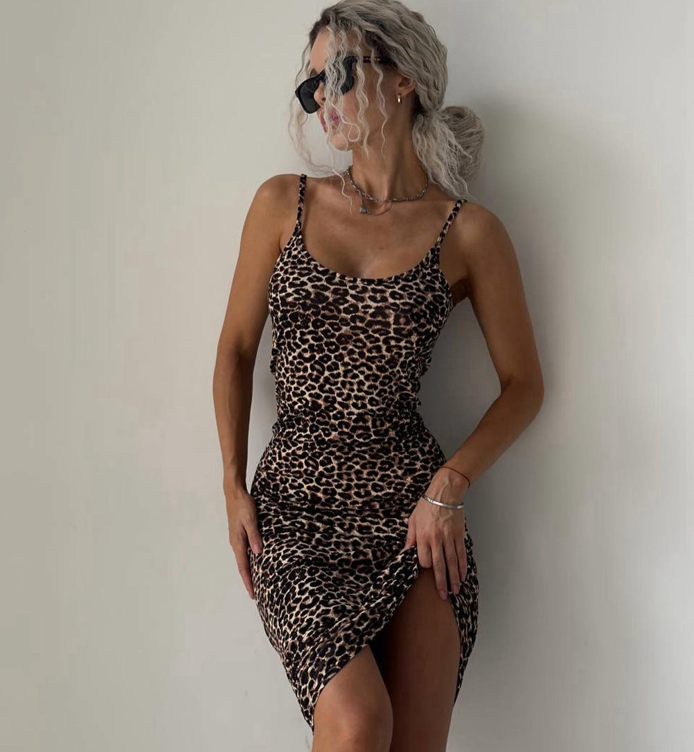 A woman with silver-gray hair and sunglasses wearing a fitted leopard-print mini dress with thin straps and a side slit, posing against a plain off-white wall.