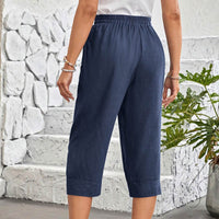 A person wearing navy blue cropped pants with an elastic waistband and cuffed hems, standing in front of a white stone wall and green plant.