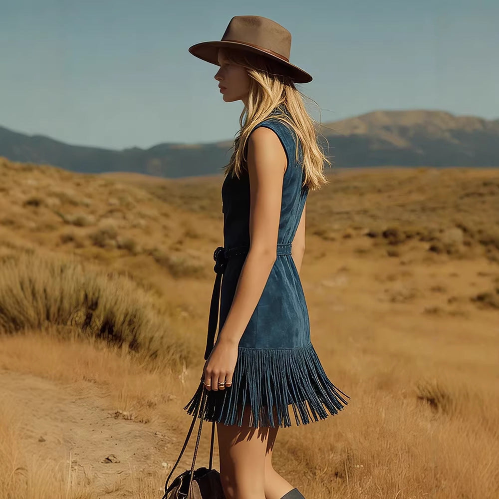 A woman stands in profile in an outdoor desert landscape wearing a sleeveless dark blue denim dress with a fringed hem and a brown wide-brim hat.