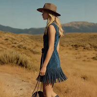 A woman stands in profile in an outdoor desert landscape wearing a sleeveless dark blue denim dress with a fringed hem and a brown wide-brim hat.
