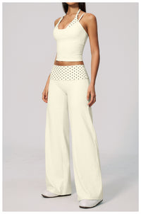 A model wearing a two-piece outfit consisting of an off-white halter crop top with cutout details and matching wide-leg pants featuring a black polka-dot patterned waistband, set against a plain light gray background.