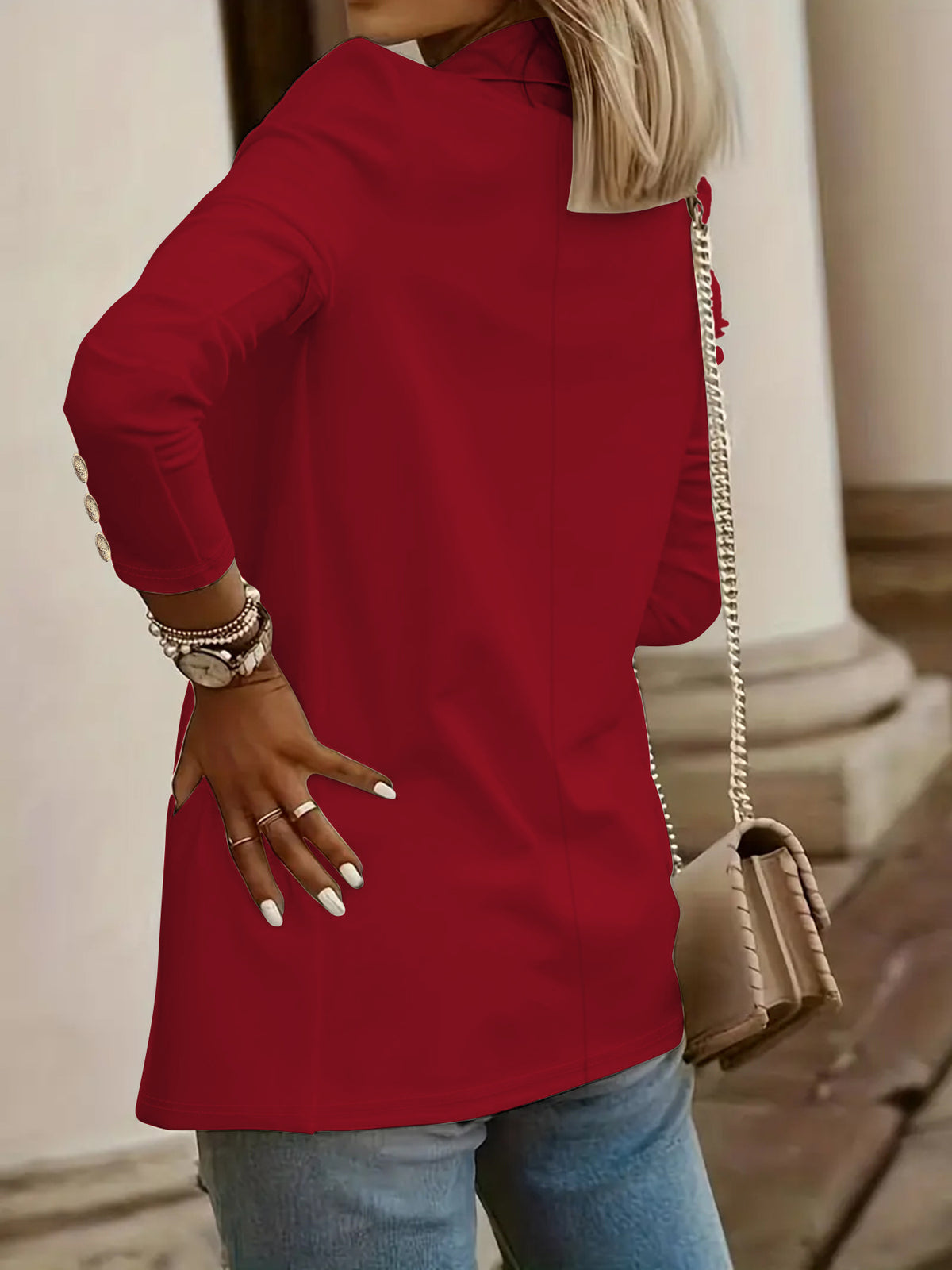 A woman wearing a bright red long-sleeve tunic top with gold button details on the cuffs, paired with blue jeans and carrying a beige shoulder bag.