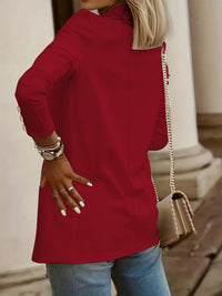A woman wearing a bright red long-sleeve tunic top with gold button details on the cuffs, paired with blue jeans and carrying a beige shoulder bag.