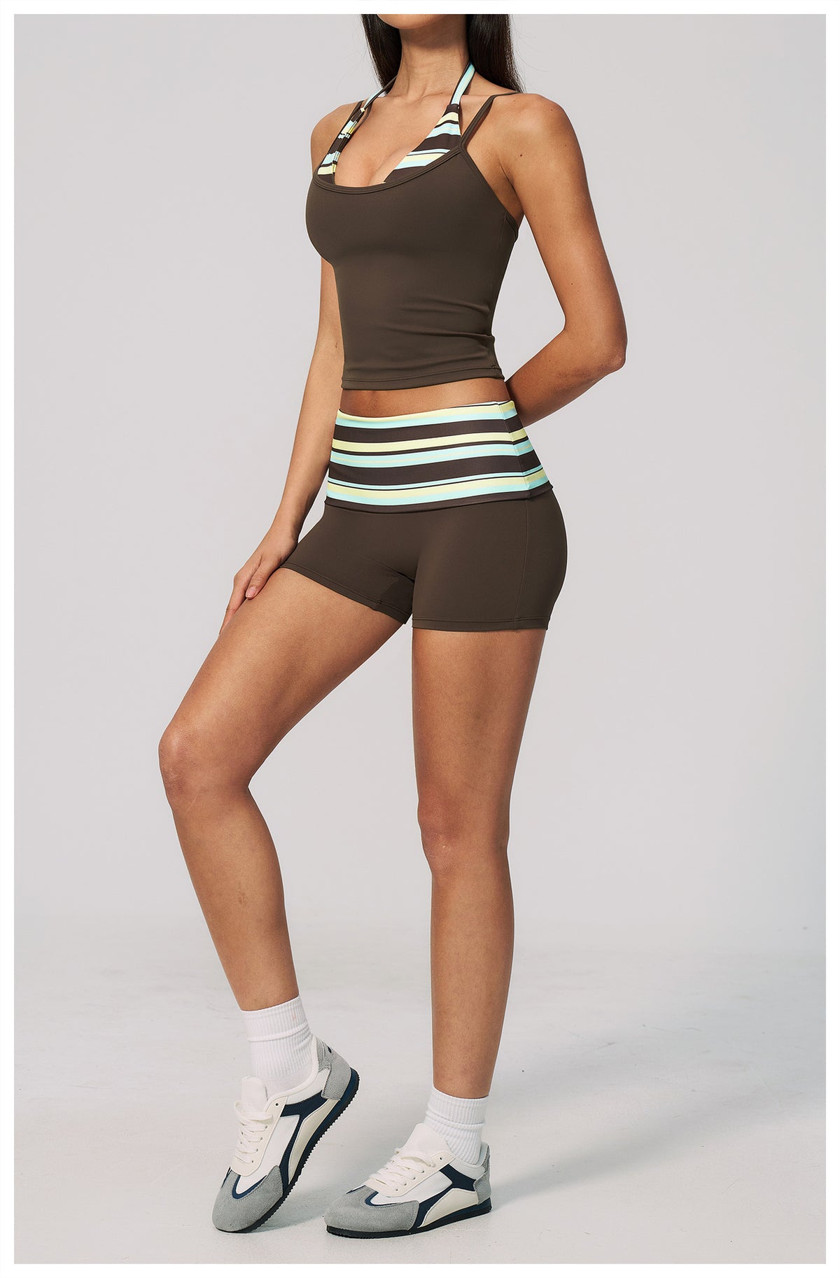 A woman modeling a matching athletic set consisting of a dark brown halter crop top and high-waisted shorts with a striped waistband in brown, white, and mint green, paired with white socks and gray sneakers, against a light gray studio background.