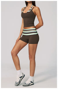 A woman modeling a matching athletic set consisting of a dark brown halter crop top and high-waisted shorts with a striped waistband in brown, white, and mint green, paired with white socks and gray sneakers, against a light gray studio background.
