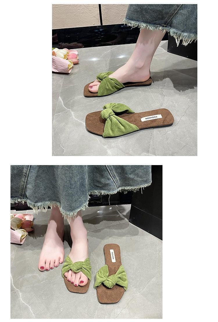A pair of women's flat slide sandals with a wide green fabric bow across the strap and a brown footbed, shown both on a model's foot and as a standalone product on a gray tiled floor.