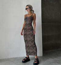 A woman modeling a sleeveless leopard-print maxi dress with thin straps, paired with black platform sandals, standing in a minimalist room with a white wall and concrete floor.