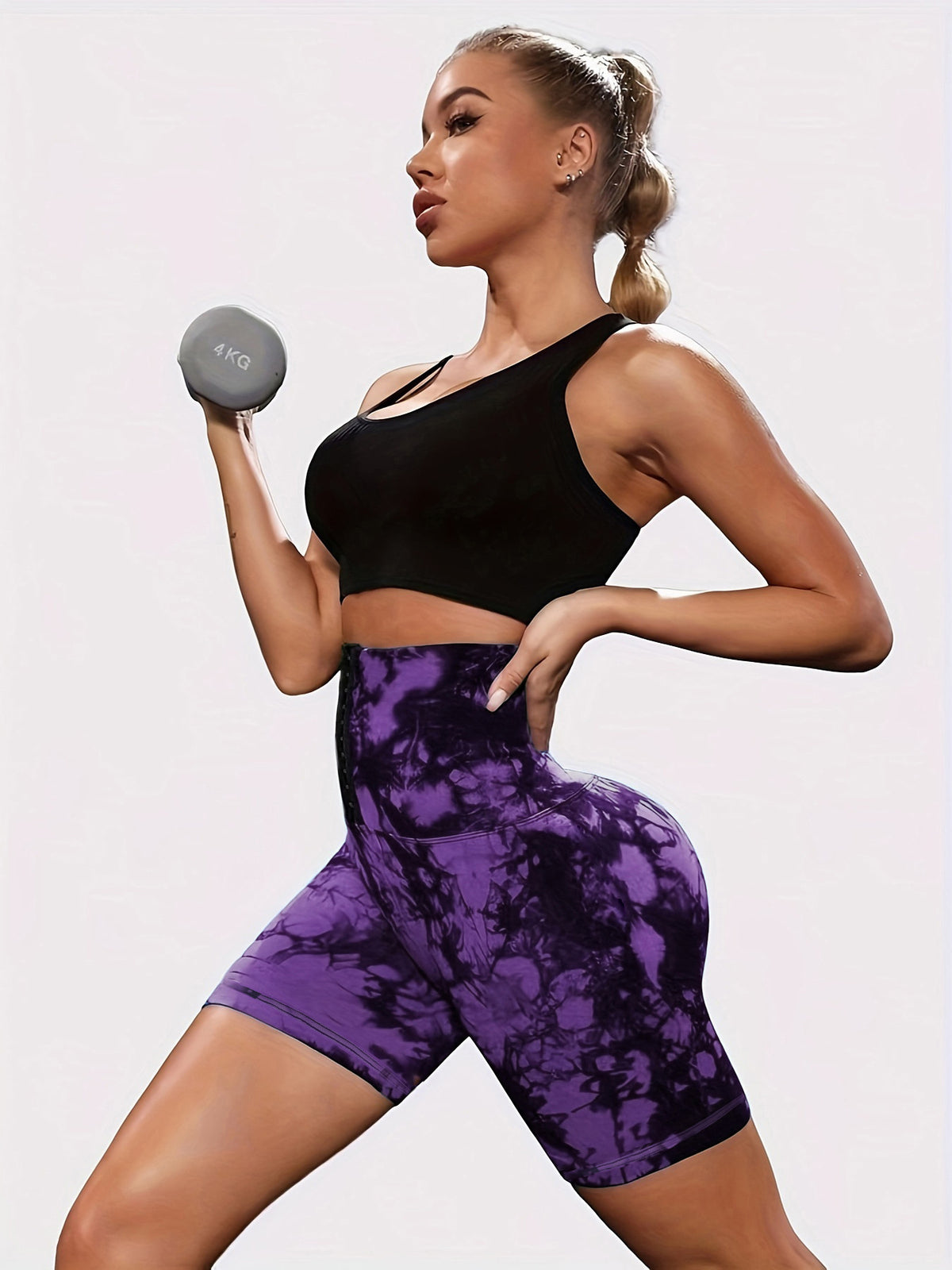 A woman in a black sports bra and purple tie-dye biker shorts holds a small gray dumbbell against a plain light background.
