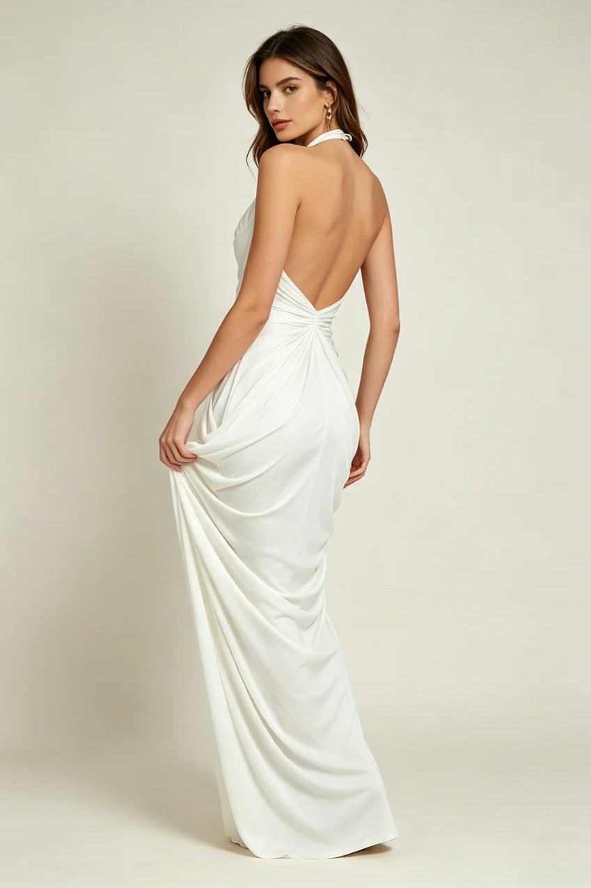 A woman modeling a floor-length white halter dress with a draped open back, shown from the back against a plain light beige studio background.