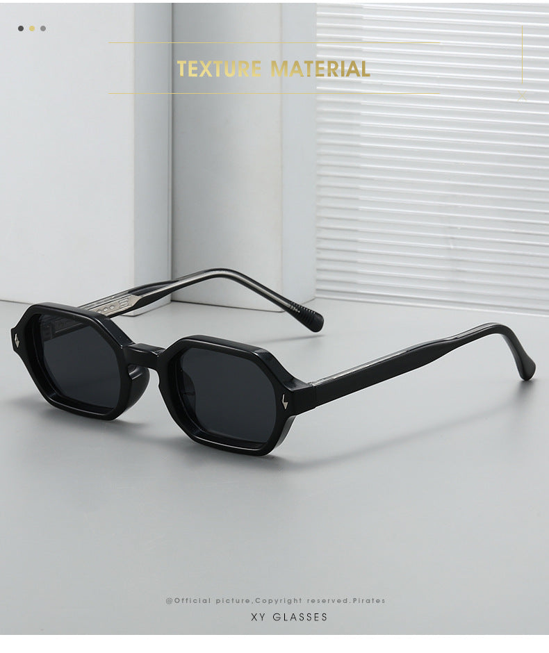 Black geometric square sunglasses with dark lenses and silver-toned metal temples, displayed on a light gray surface with a white window-blind background. Text overlays read 'TEXTURE MATERIAL' and 'XY GLASSES'.