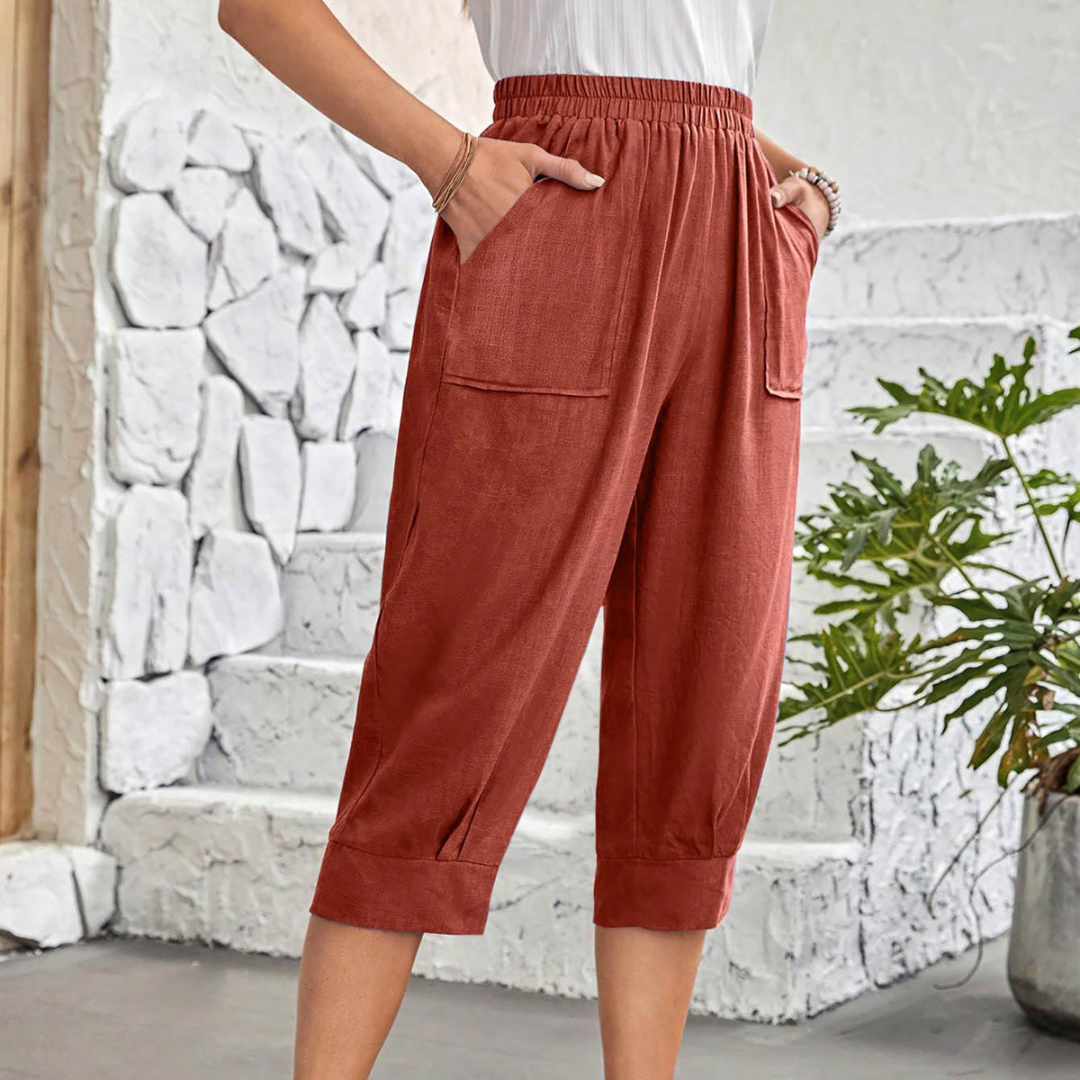 A person wearing loose, wide-leg cropped pants in a muted terracotta or rust color, standing indoors against a white stone wall with a green plant at the side.