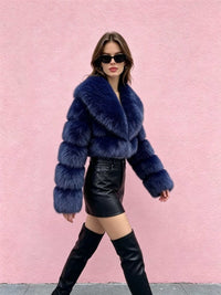 A model wearing a voluminous dark blue faux fur cropped jacket, black leather mini skirt, and black thigh-high boots, posing against a textured pink wall.