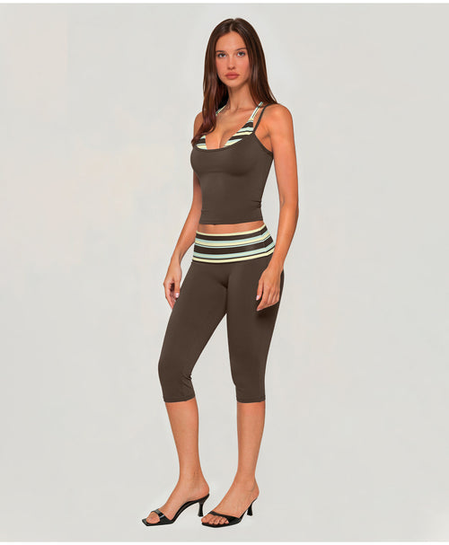 A woman modeling a matching activewear set consisting of a dark brown scoop-neck tank top and high-waisted capri leggings, both featuring a striped waistband in green, white, and brown, paired with black heeled sandals against a light gray background.