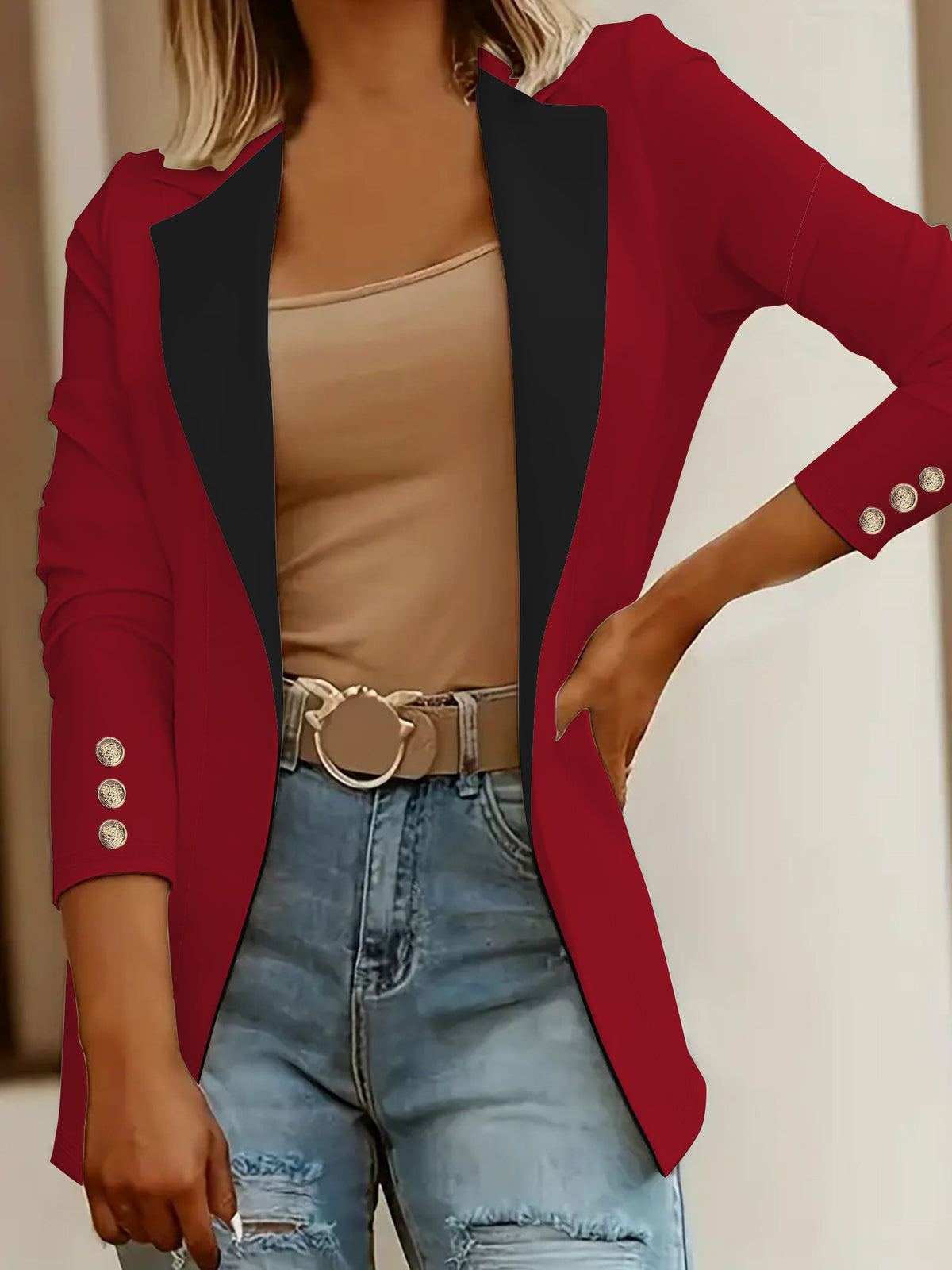 A woman wearing a red blazer with black lapels, styled over a beige tank top and light blue distressed jeans with a decorative belt.