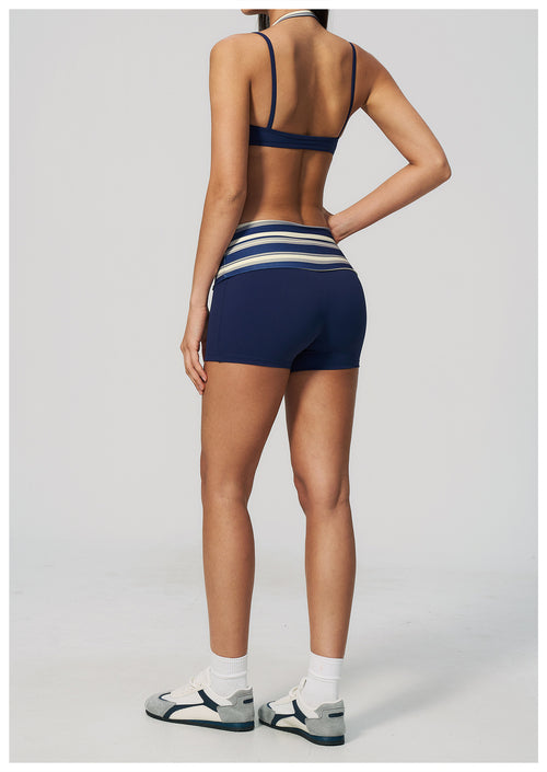 Rear view of a model wearing navy blue athletic shorts with white and light blue horizontal stripes at the waistband, paired with a matching navy sports bra and white sneakers, against a light gray studio background.