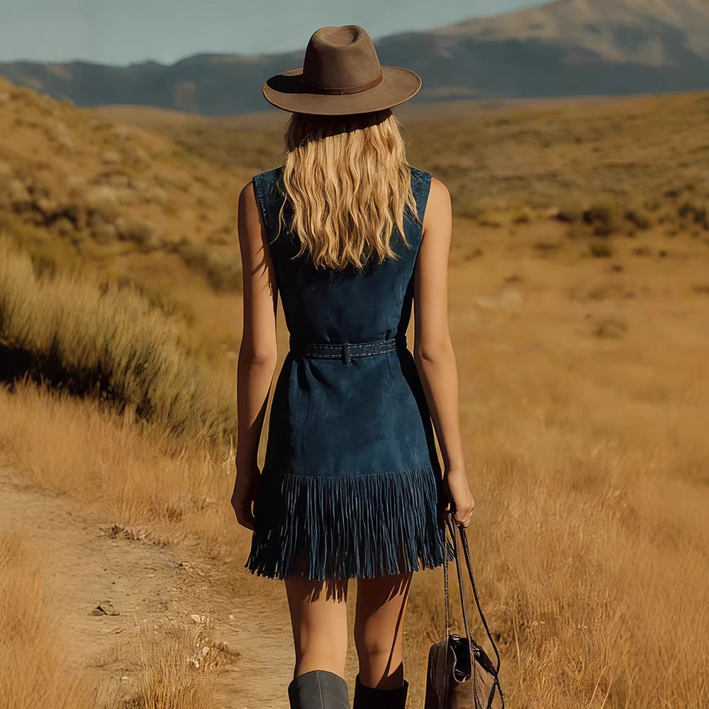 A woman seen from behind walking on a dirt path in a dry grassy landscape, wearing a sleeveless dark blue denim dress with fringe, a brown wide-brim hat, and carrying a dark shoulder bag.