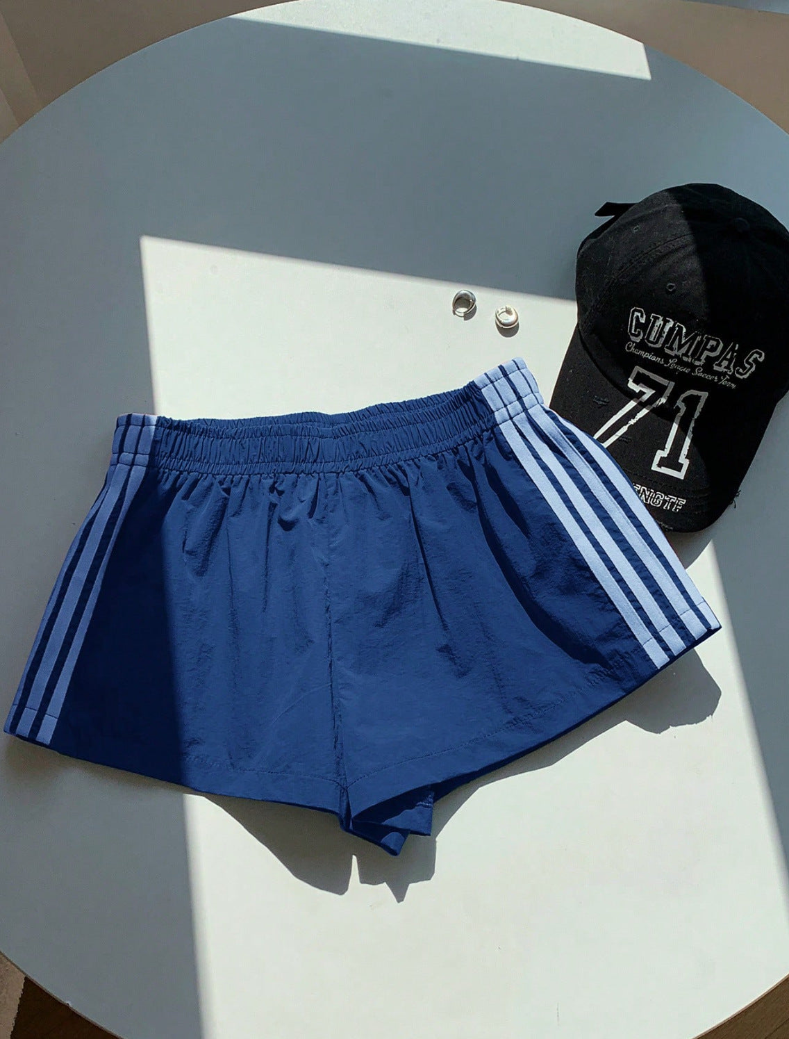A pair of royal blue athletic shorts with white side stripes, laid flat on a white circular table next to a black baseball cap.