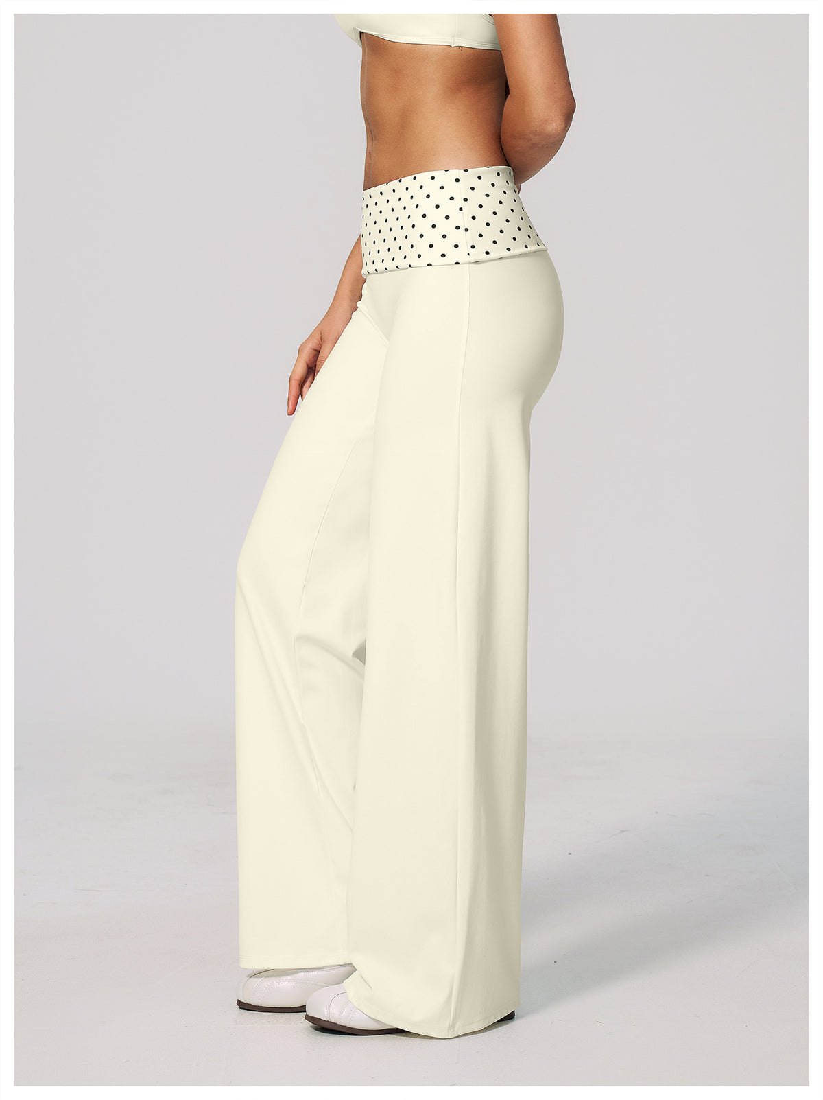 A model is shown from the waist down wearing high-waisted, wide-leg pants in cream with a contrasting white waistband featuring black polka dots.