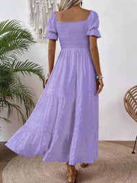 A woman wearing a lavender purple tiered maxi dress with puff sleeves and a smocked bodice, shown from the back in a styled indoor setting.