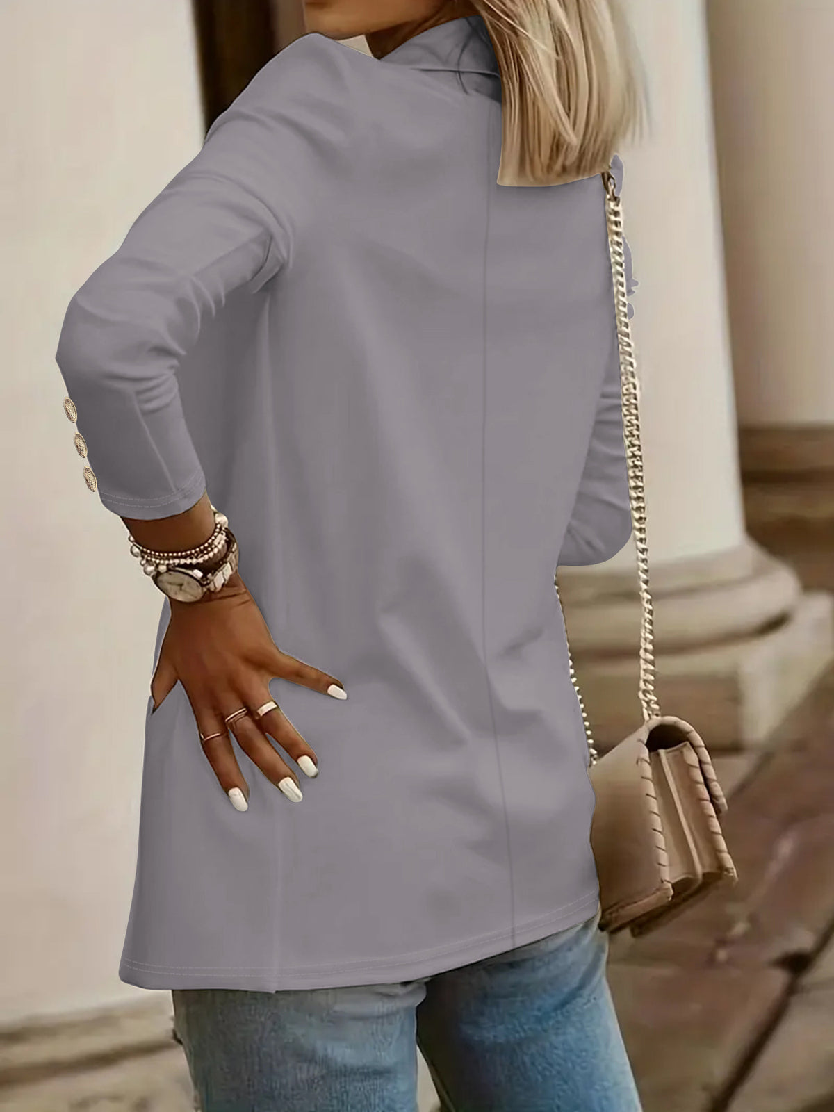 A woman wearing a light gray long-sleeve tunic top with gold button details on the cuffs, paired with blue jeans and a beige chain strap handbag.