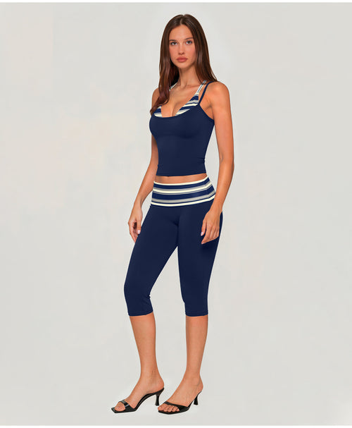 A woman modeling a matching navy blue activewear set consisting of a scoop-neck tank top and high-waisted capri leggings, both featuring white and navy striped details, set against a plain light gray background.