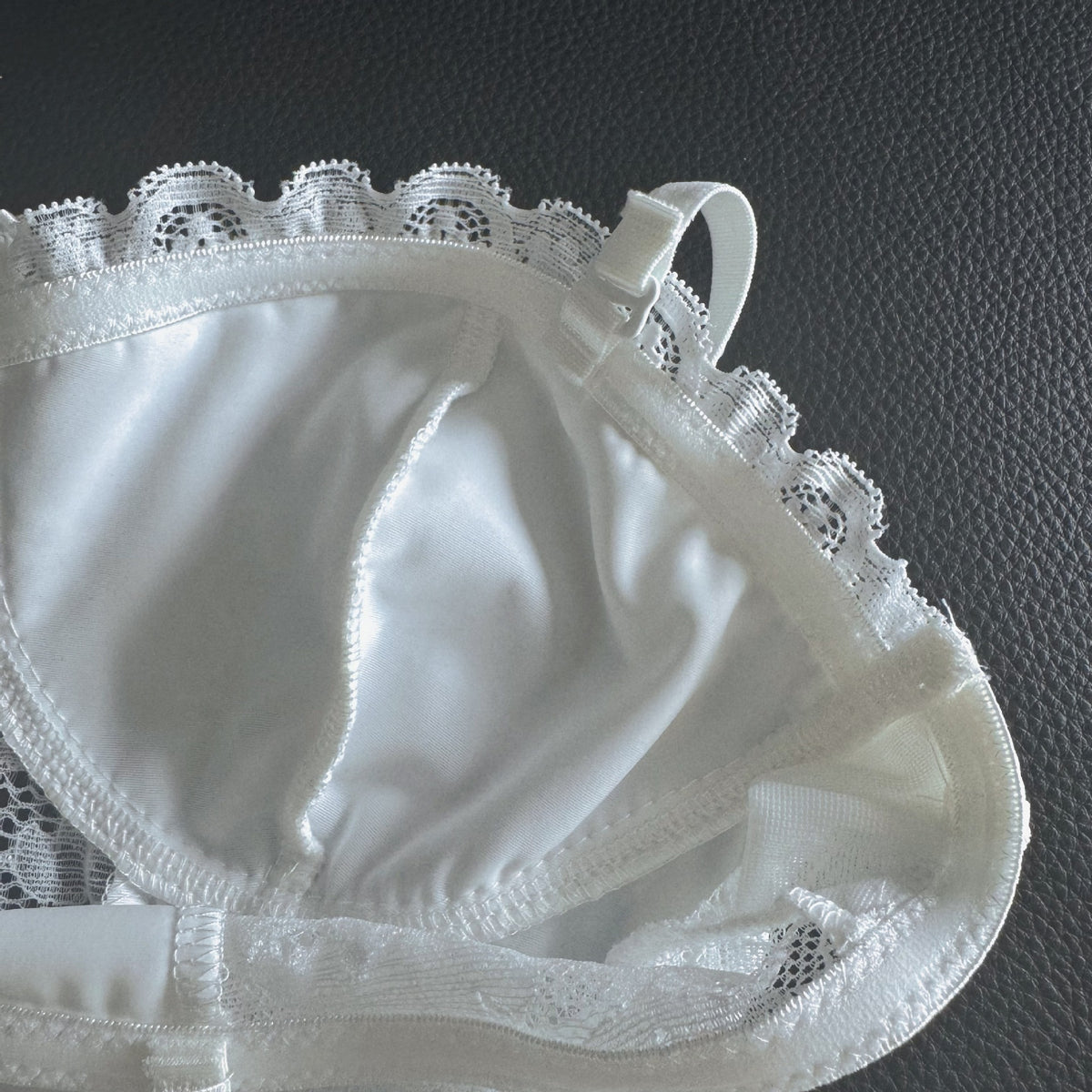 White lace bra cup with satin-like fabric and scalloped lace trim, photographed close up on a dark textured surface.