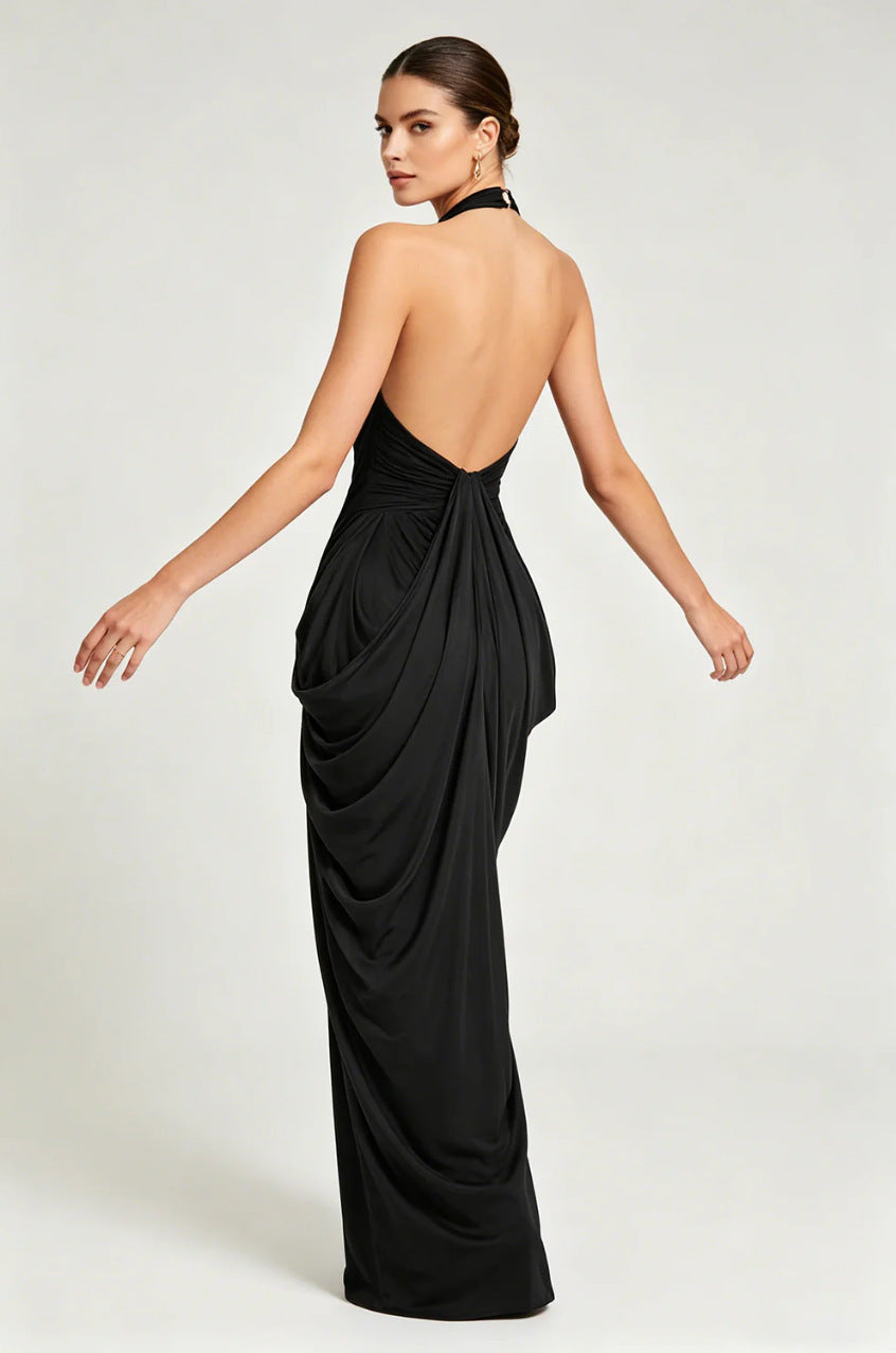 A woman modeling a floor-length black halter gown from the back, featuring a draped open back and gathered skirt, against a light gray studio background.